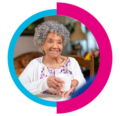 Older woman in purple floral shirt holding tea mug while chatting with home health aide during a home visit.