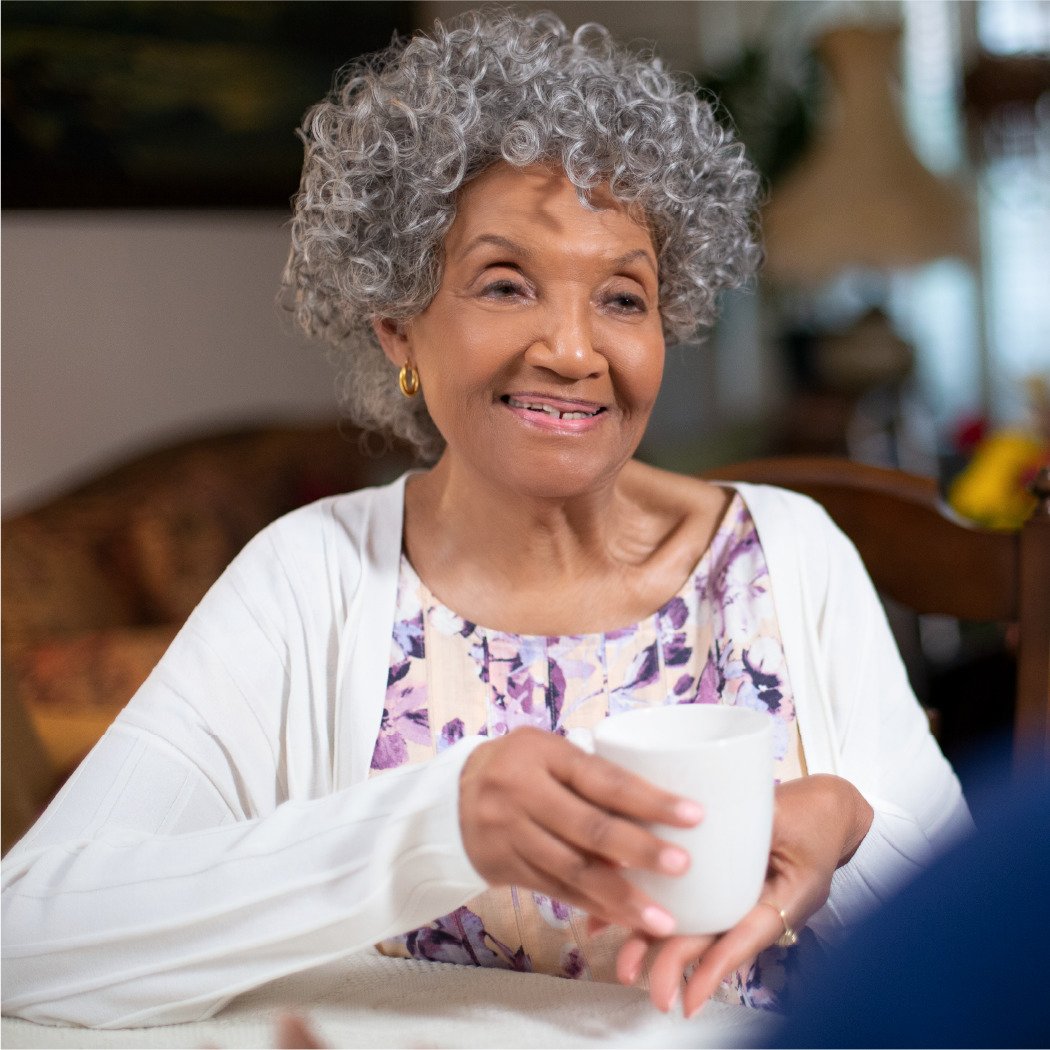 Older woman in purple floral shirt holding tea mug while chatting with home health aide during a home visit.
